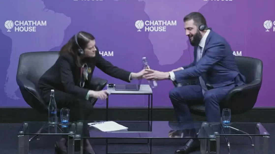 Syrian President Ahmed al-Sharaa at a Chatham House event, reaching to hand back a water bottle he could not open during an interview.
