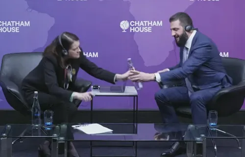 Syrian President Ahmed al-Sharaa at a Chatham House event, reaching to hand back a water bottle he could not open during an interview.